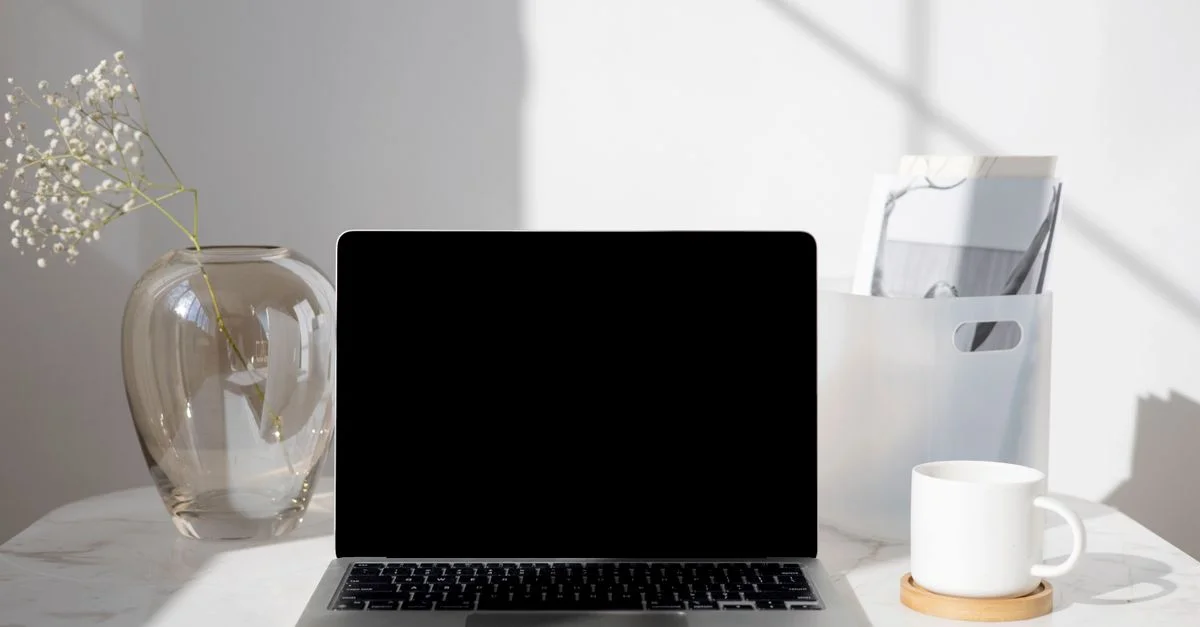 Un bureau propre et moderne avec un ordinateur portable, une tasse de café, un vase et des documents sous une lumière du jour éclatante.